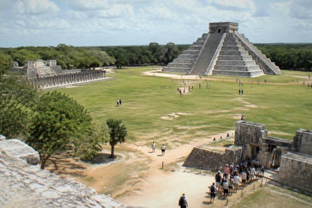 A wide grassy plaza bordered by grey stone ruins under a partly cloudy sky