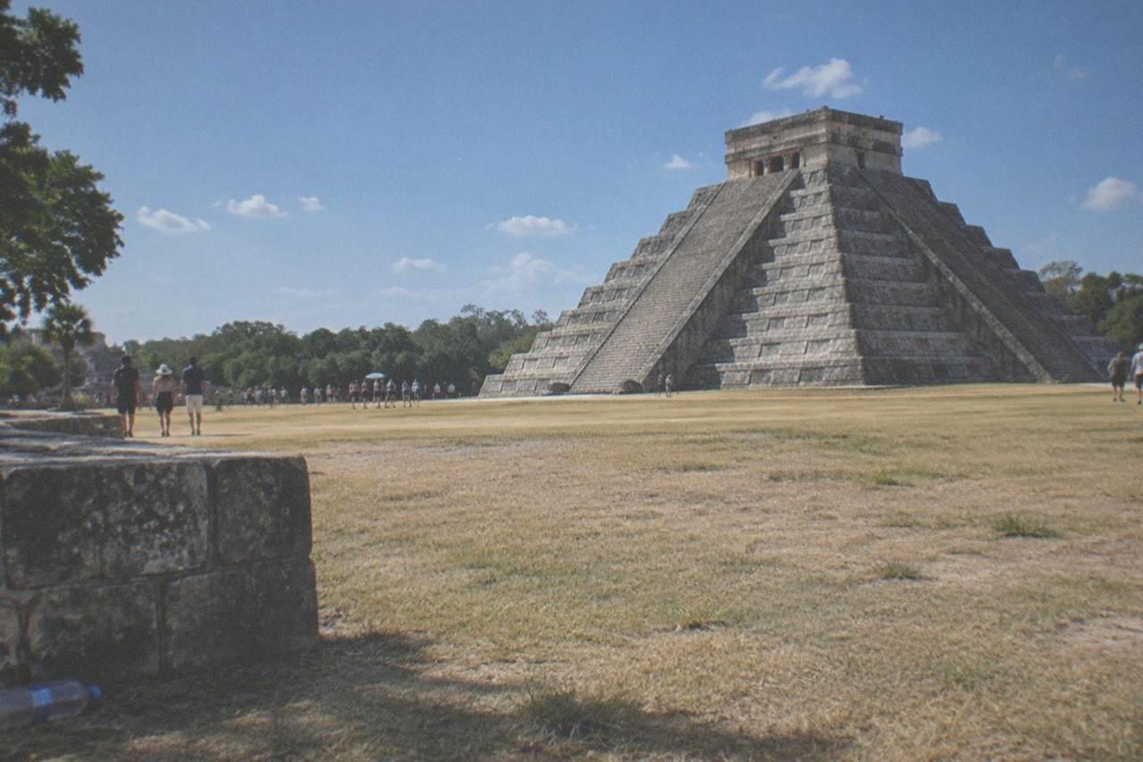 The stepped pyramid of El Castillo rising above open grass and low stone platforms under a bright sky