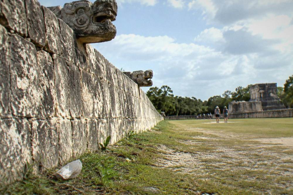 A long stone wall with carved serpent heads at the edge of the great ball court under scattered clouds