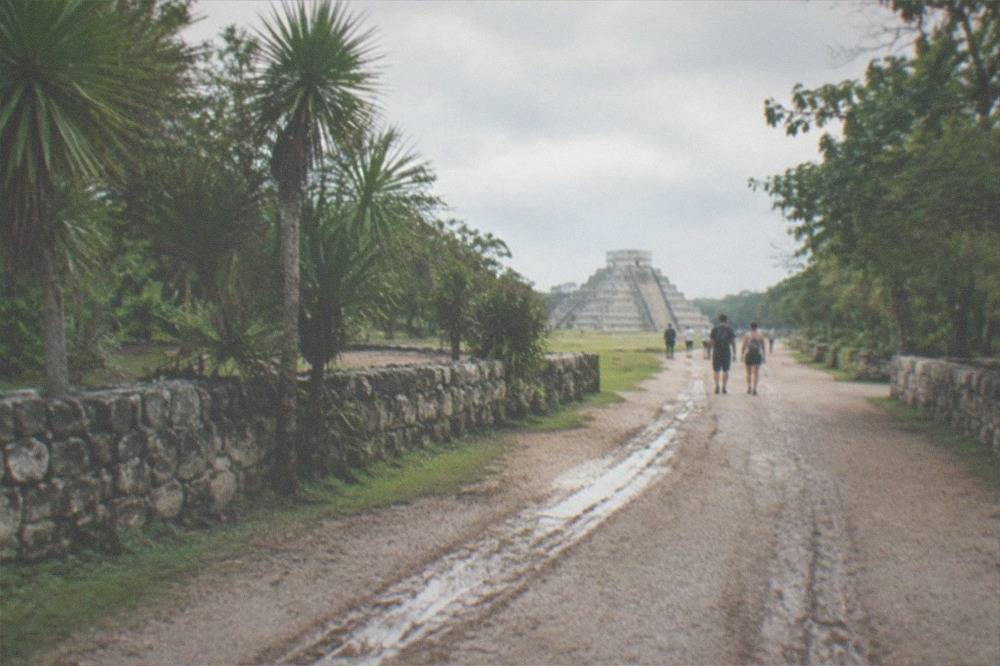 A wide gravel path leading toward the distant pyramid with low stone walls and trees on either side