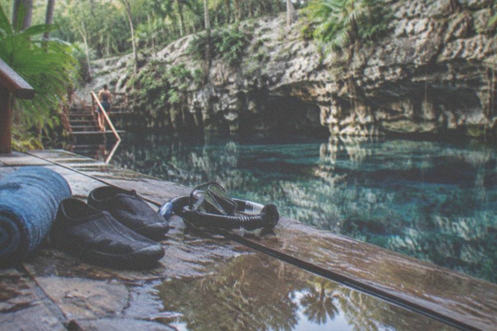 Water shoes, towel, and snorkel mask set out on a wooden cenote deck beside still blue water