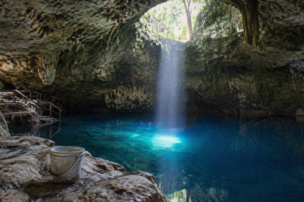 Sunlight beams falling through a round opening in a cave ceiling onto deep blue water