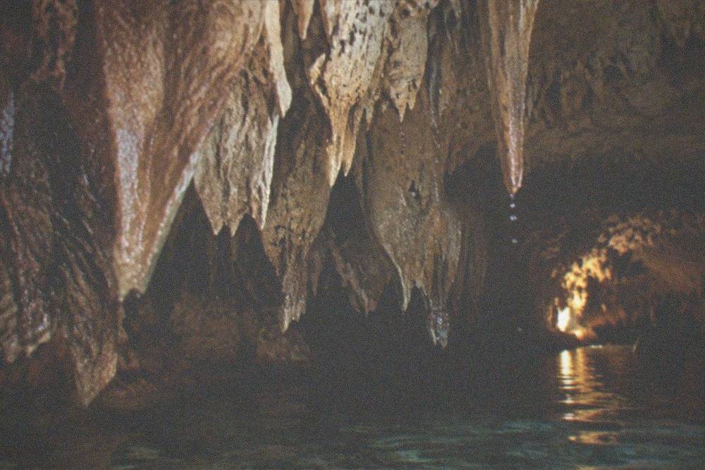 Stalactites hanging from a dim cave ceiling above a still underground pool lit by a guide lamp