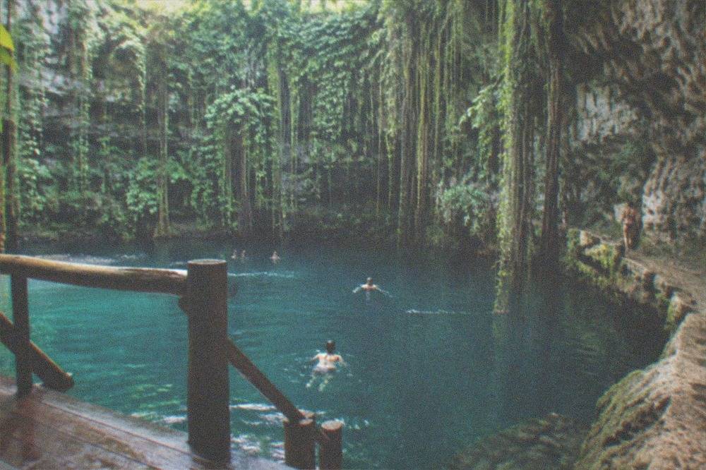 Turquoise open cenote surrounded by rock walls and hanging vines with a wooden platform along one edge