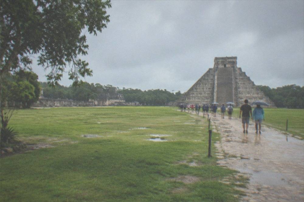 Long grassy plaza at Chichen Itza with the pyramid in the distance and visitors walking along a stone path
