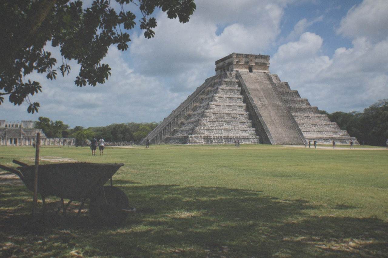 The stepped limestone pyramid of El Castillo rising above green lawn at Chichen Itza under a partly cloudy sky
