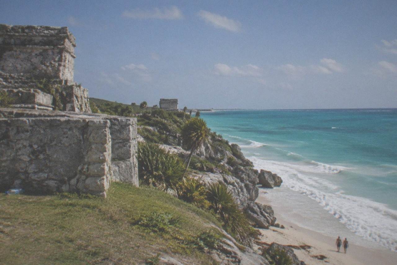 Ancient stone ruins on a grassy cliff above a turquoise Caribbean sea with waves breaking on sand below