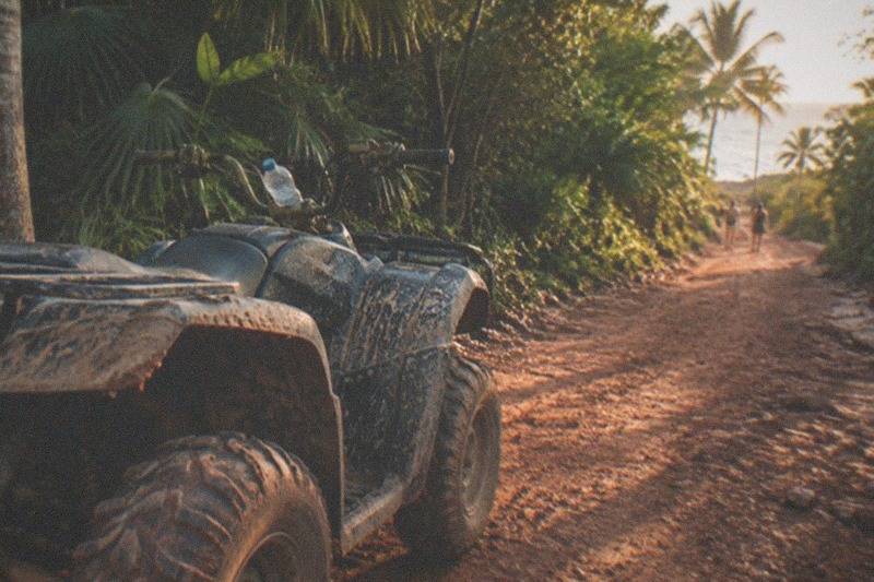 A mud-splashed ATV buggy parked on a red dirt jungle trail near Playa del Carmen, dense tropical foliage around it, no people.