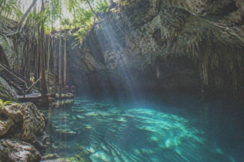 A clear freshwater cenote cavern near Playa del Carmen, sunlight beams through a stone opening onto turquoise water, limestone stalactites and jungle roots at the edge, no people.