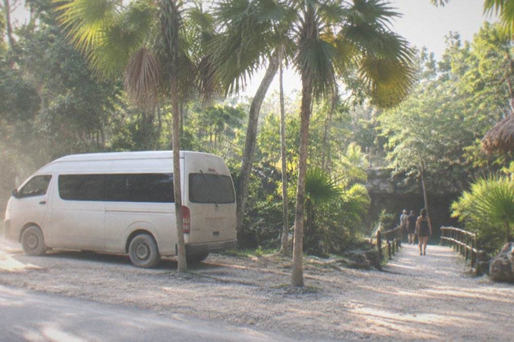 Shared van parked beside a jungle highway with a gravel path leading toward cenote entrances