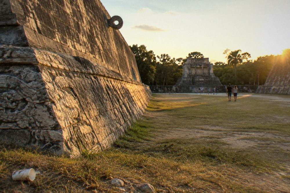 Open grassy plaza at Chichen Itza with the carved stone wall of the ballcourt and visitors on paths in the distance
