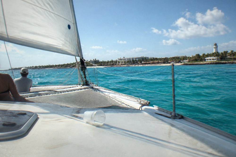 Catamaran with sails up crossing open turquoise water toward a palm-lined island