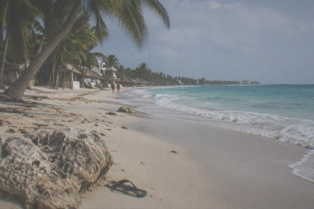 A wide empty Caribbean beach with palm trees and gentle waves under a hazy sky