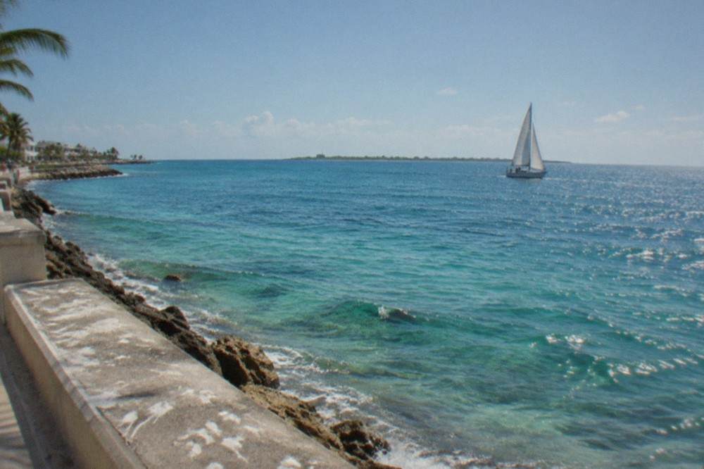Open Caribbean water with a distant island shoreline and a sailboat under a clear sky.
