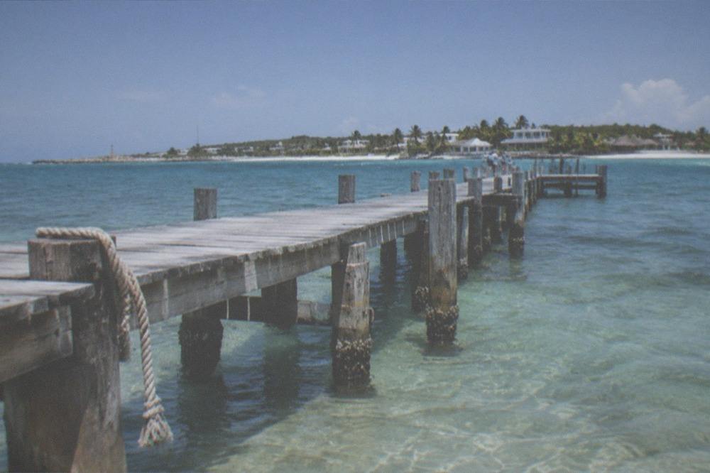 Wooden pier extending over clear shallow water toward a distant island silhouette at midday.