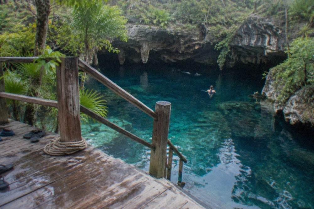 Clear blue cenote water seen from a wooden platform with jungle foliage around the opening
