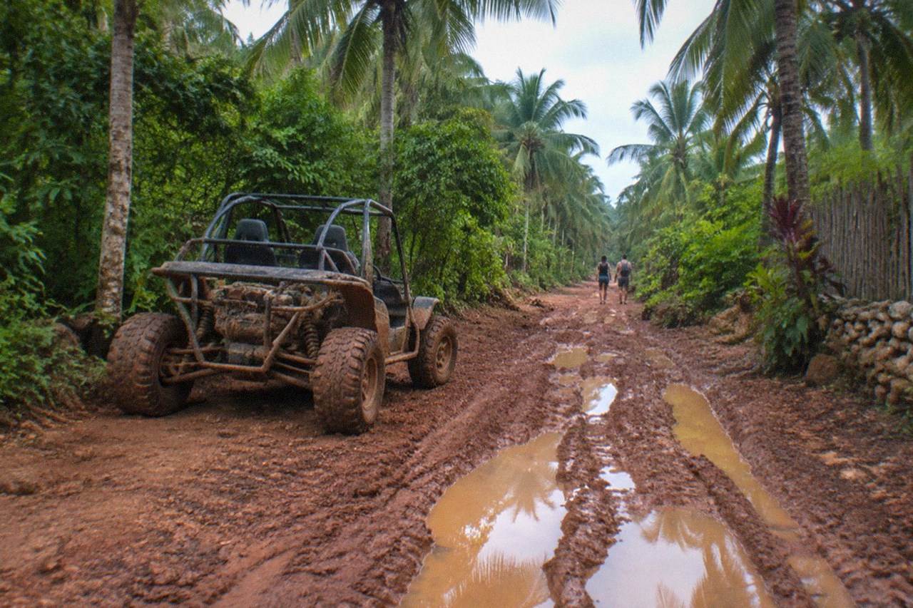 Wide establishing view. A mud-splashed ATV buggy parked on a red dirt jungle trail near Playa del Carmen, dense tropical foliage around it, no people.