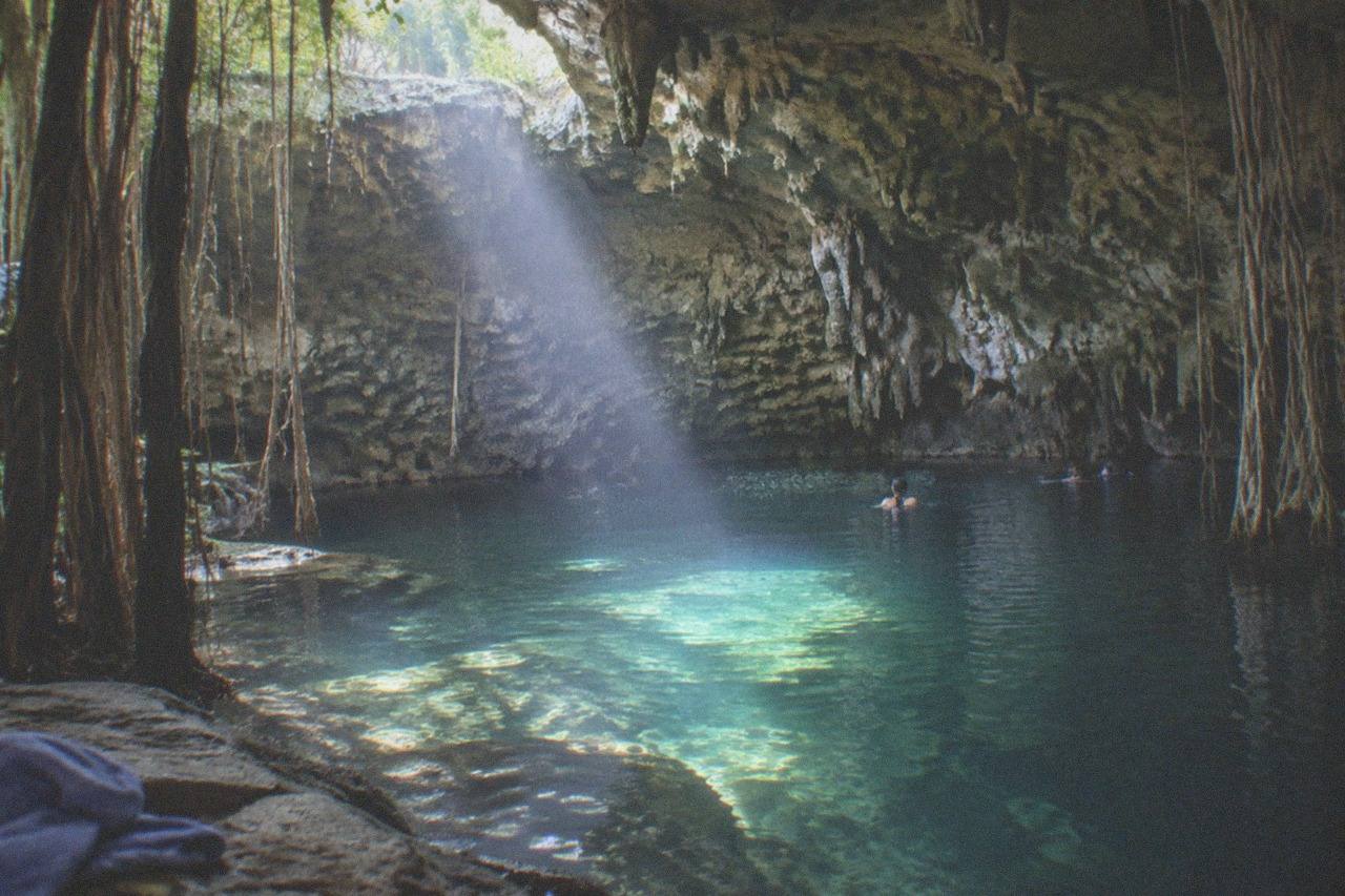 Wide establishing view. A clear freshwater cenote cavern near Playa del Carmen, sunlight beams through a stone opening onto turquoise water, limestone stalactites and jungle roots at the edge, no people.