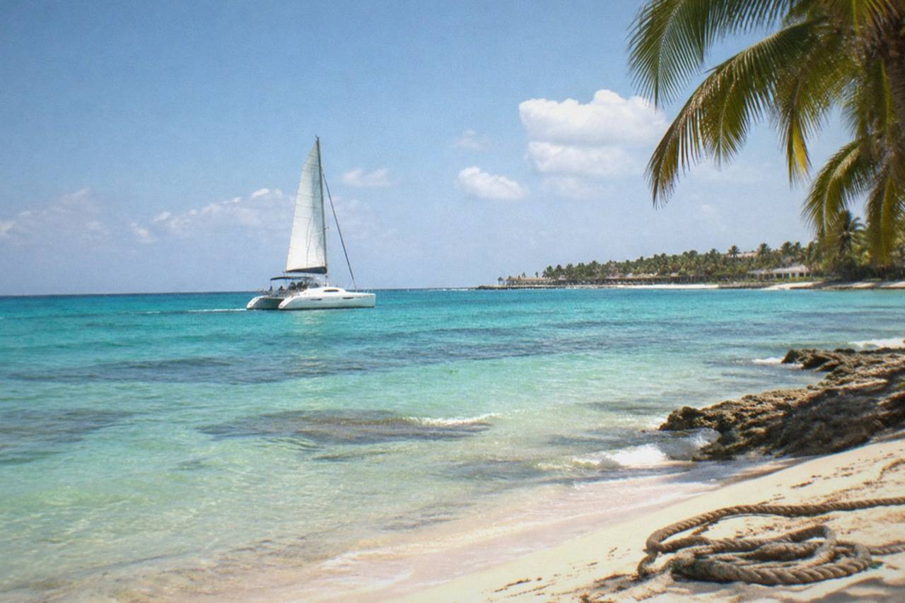 Wide establishing view. A Caribbean catamaran sailing on turquoise water off Cozumel, white sail and wake, coral-reef colors visible below the surface, no people.